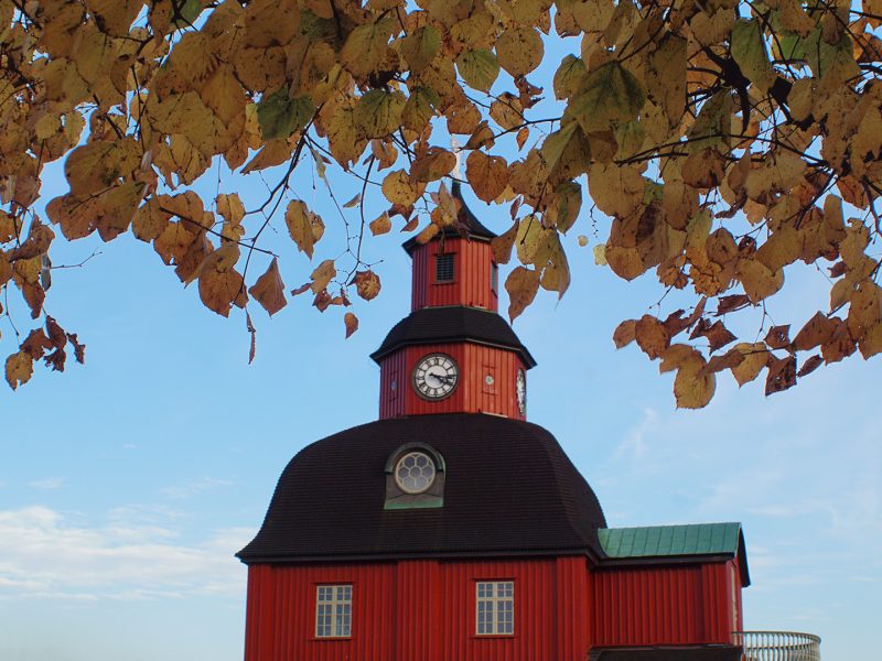 The image shows a bright red town hall in Lidköping, Sweden, with a clock tower, viewed through the frame of golden autumn leaves against a clear blue sky.