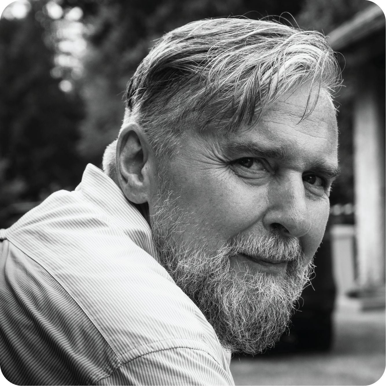 Black and white photograph of an older man with a gray beard and hair, looking over his shoulder. He is wearing a striped shirt and is outdoors with a blurred background of trees and a building. The edges of the image are rounded. How is working about part-time photographer